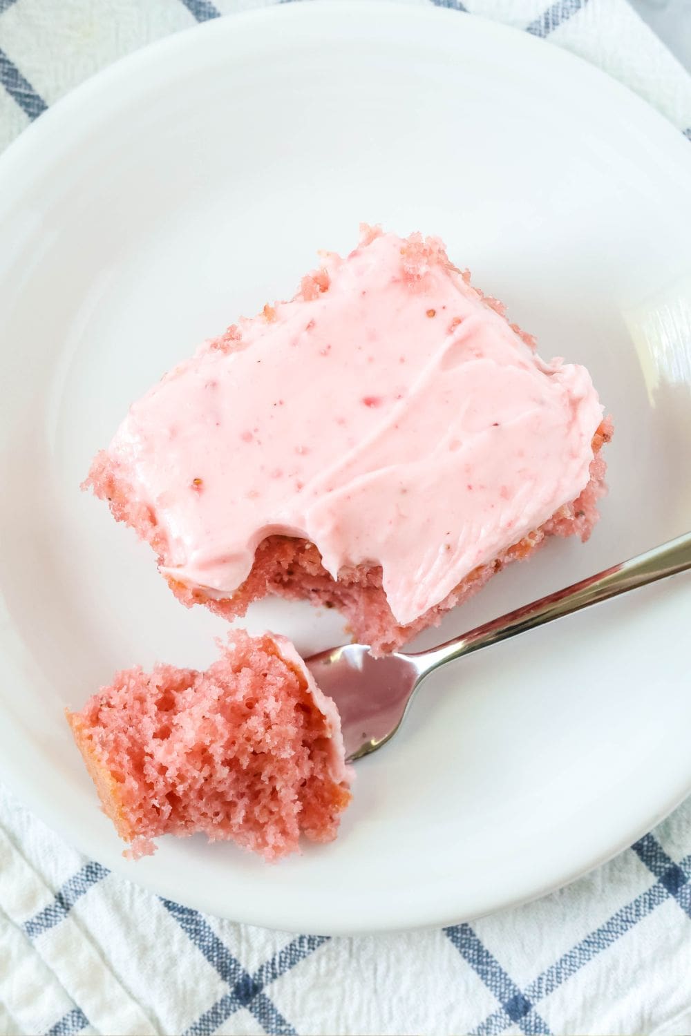 overhead view of a slice of strawberry cake with strawberry frosting, served on a white plate. A fork rests on the plate with a bite of cake on it.