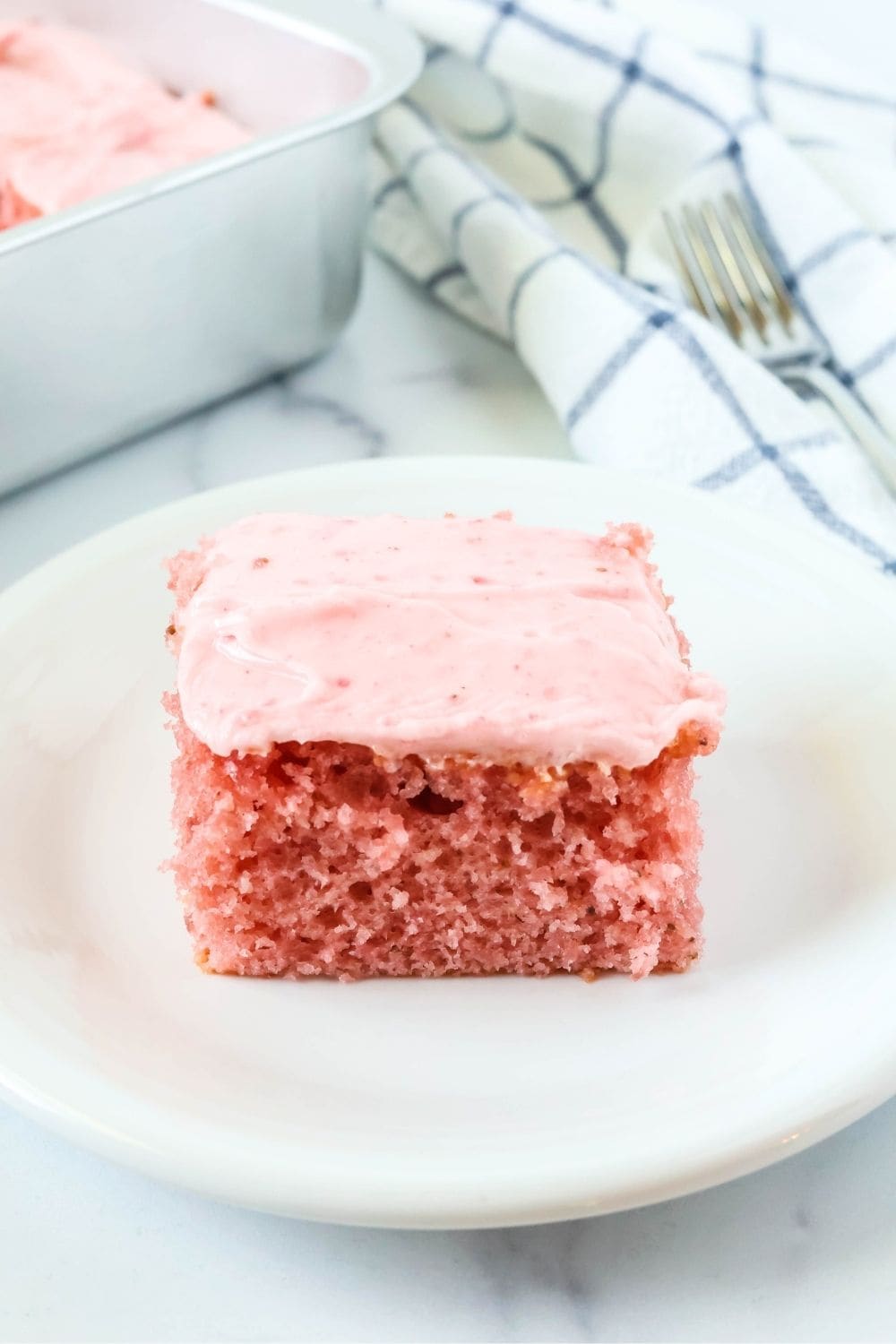 Square slice of frosted strawberry Jello cake is served on a white plate. A fork and the cake pan are in the background.