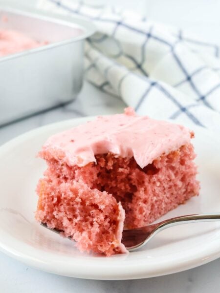 Strawberry sheet cake slice topped with strawberry cream cheese frosting is served on a white plate; a fork has removed a bite of the cake.