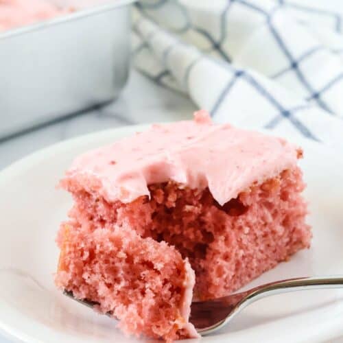Strawberry sheet cake slice topped with strawberry cream cheese frosting is served on a white plate; a fork has removed a bite of the cake.
