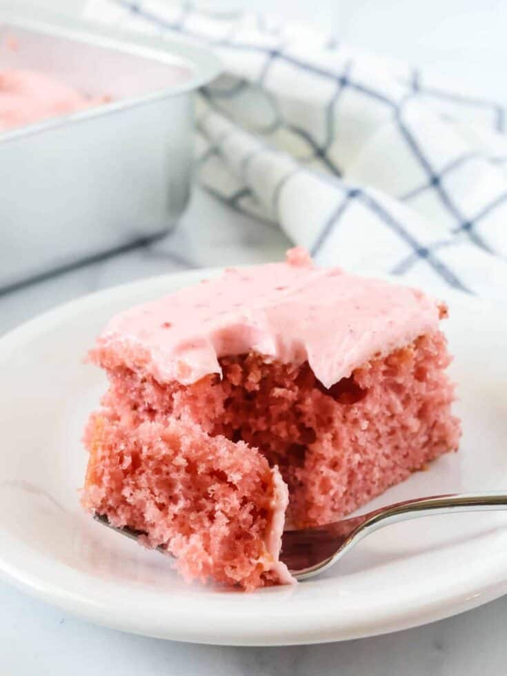 Strawberry sheet cake slice topped with strawberry cream cheese frosting is served on a white plate; a fork has removed a bite of the cake.