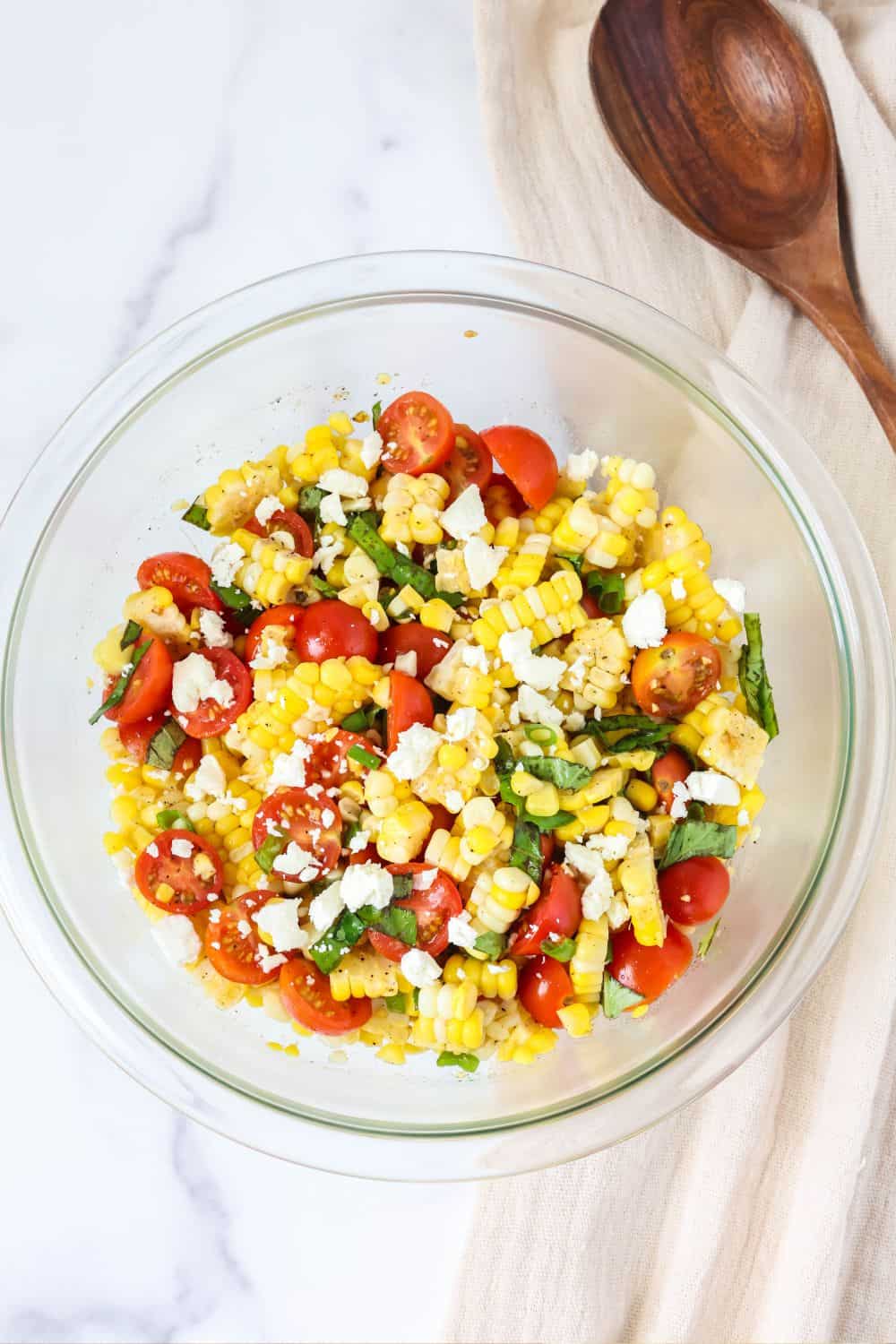 overhead view of a glass bowl of tomato basil corn salad with feta cheese. A wooden serving spoon is next to the bowl.