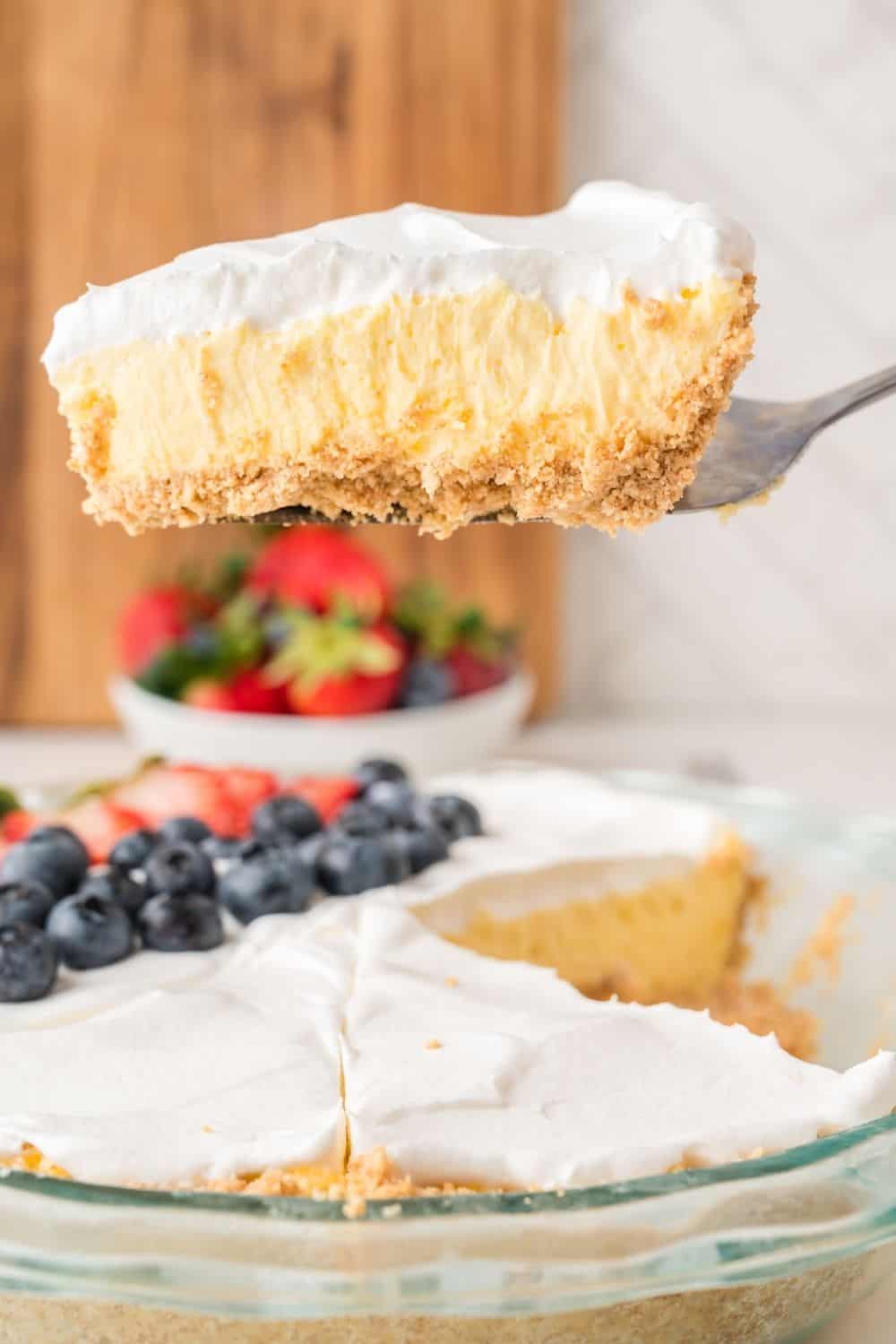 A pie server lifts a slice of vanilla cream pie from the pie dish, showcasing the creamy vanilla pudding pie filling topped with whipped cream.