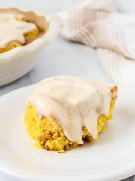 Quick pumpkin cinnamon roll topped with cinnamon cream cheese frosting, served on a white plate. A pan of remaining pumpkin cinnamon rolls is in the background.
