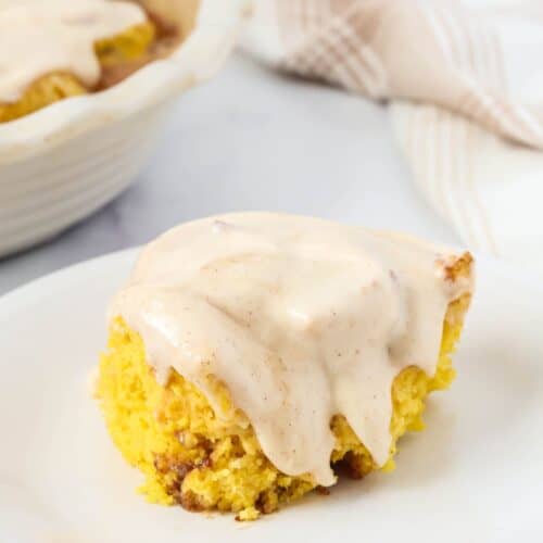 Quick pumpkin cinnamon roll topped with cinnamon cream cheese frosting, served on a white plate. A pan of remaining pumpkin cinnamon rolls is in the background.