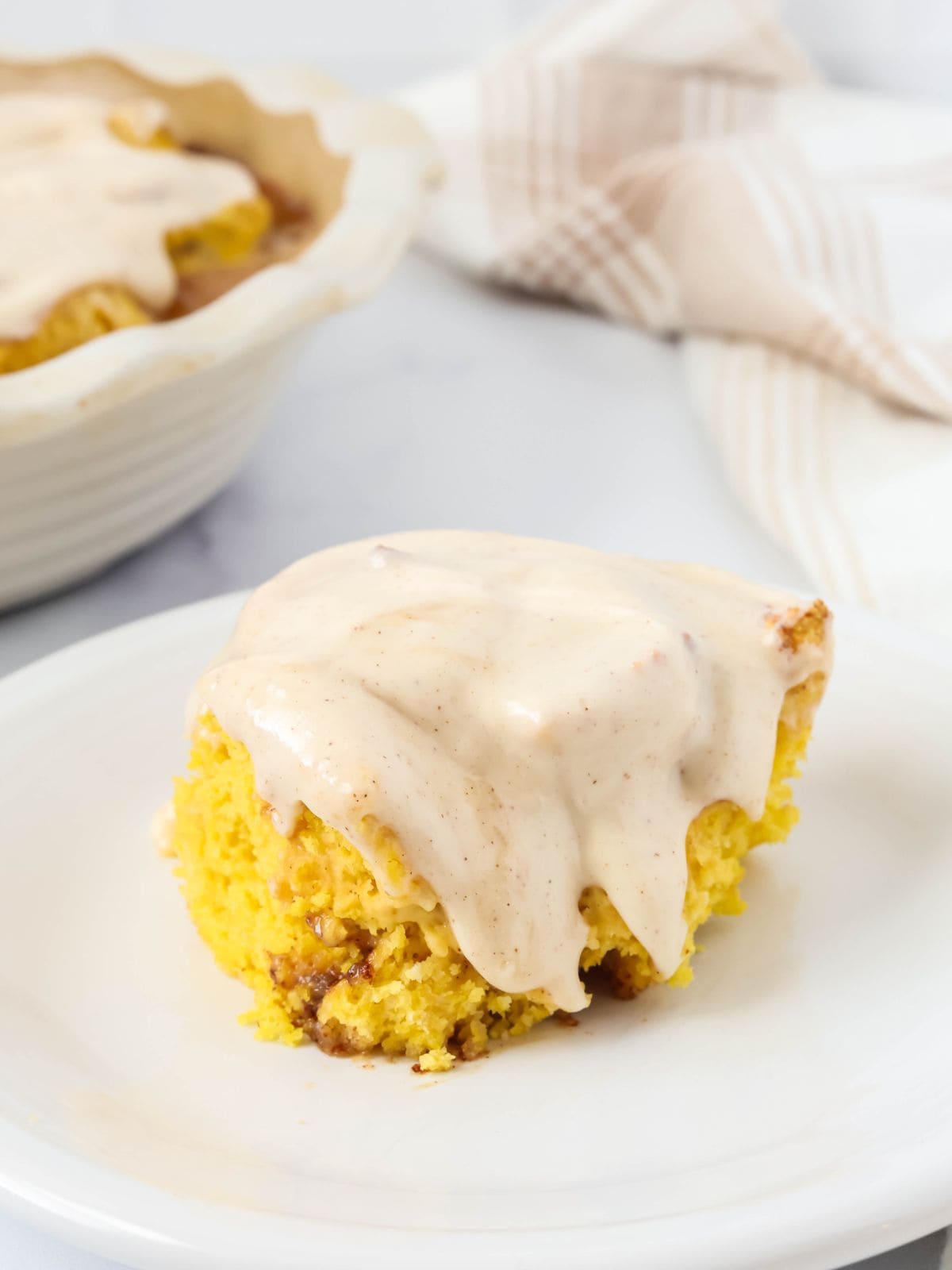 Quick pumpkin cinnamon roll topped with cinnamon cream cheese frosting, served on a white plate. A pan of remaining pumpkin cinnamon rolls is in the background.