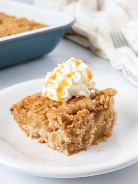 Slice of apple cake made with cake mix and pie filling, served on a white plate, topped with whipped cream and caramel drizzle.