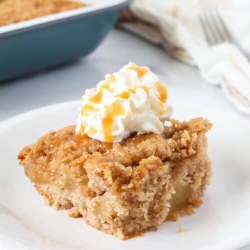 Slice of apple cake made with cake mix and pie filling, served on a white plate, topped with whipped cream and caramel drizzle.