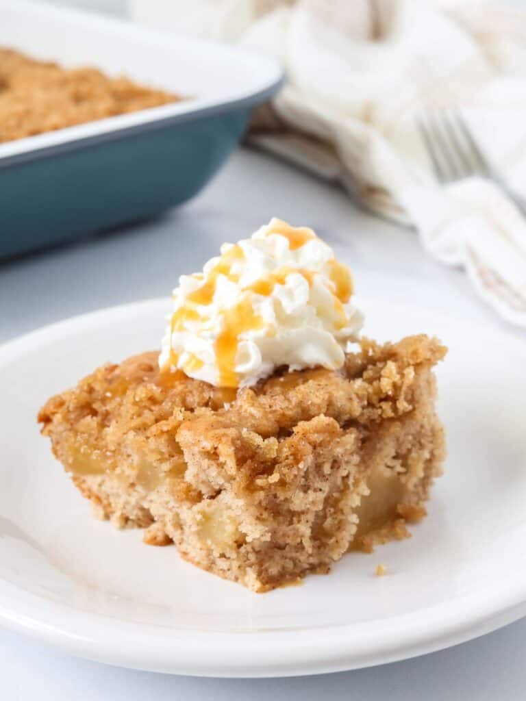 Slice of apple cake made with cake mix and pie filling, served on a white plate, topped with whipped cream and caramel drizzle.