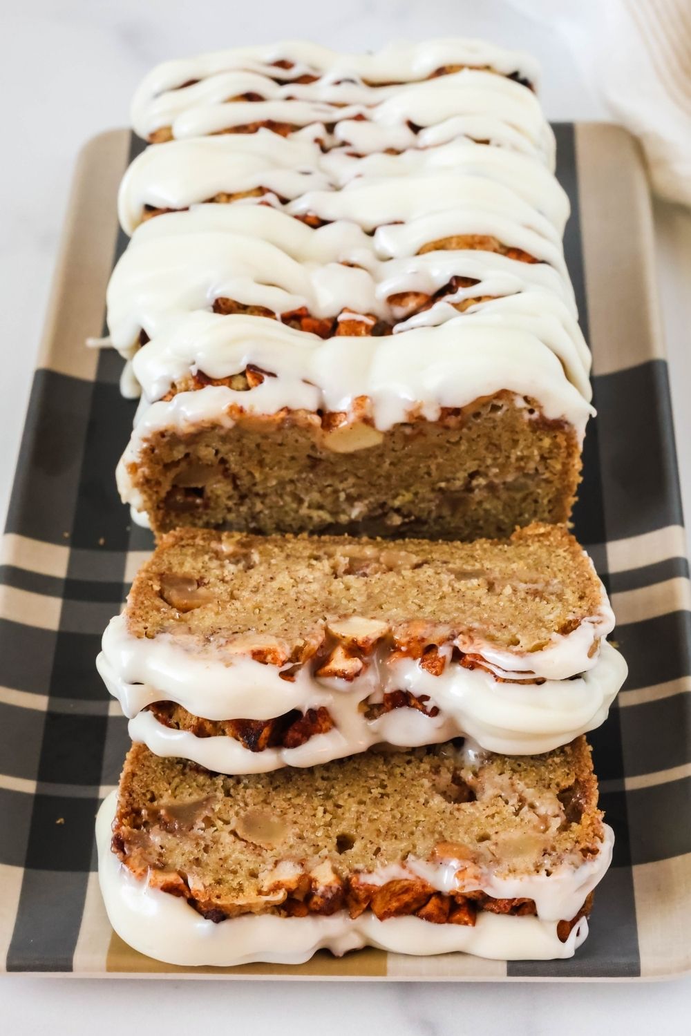 Partially sliced loaf of apple fritter bread with icing, served on a plaid platter.