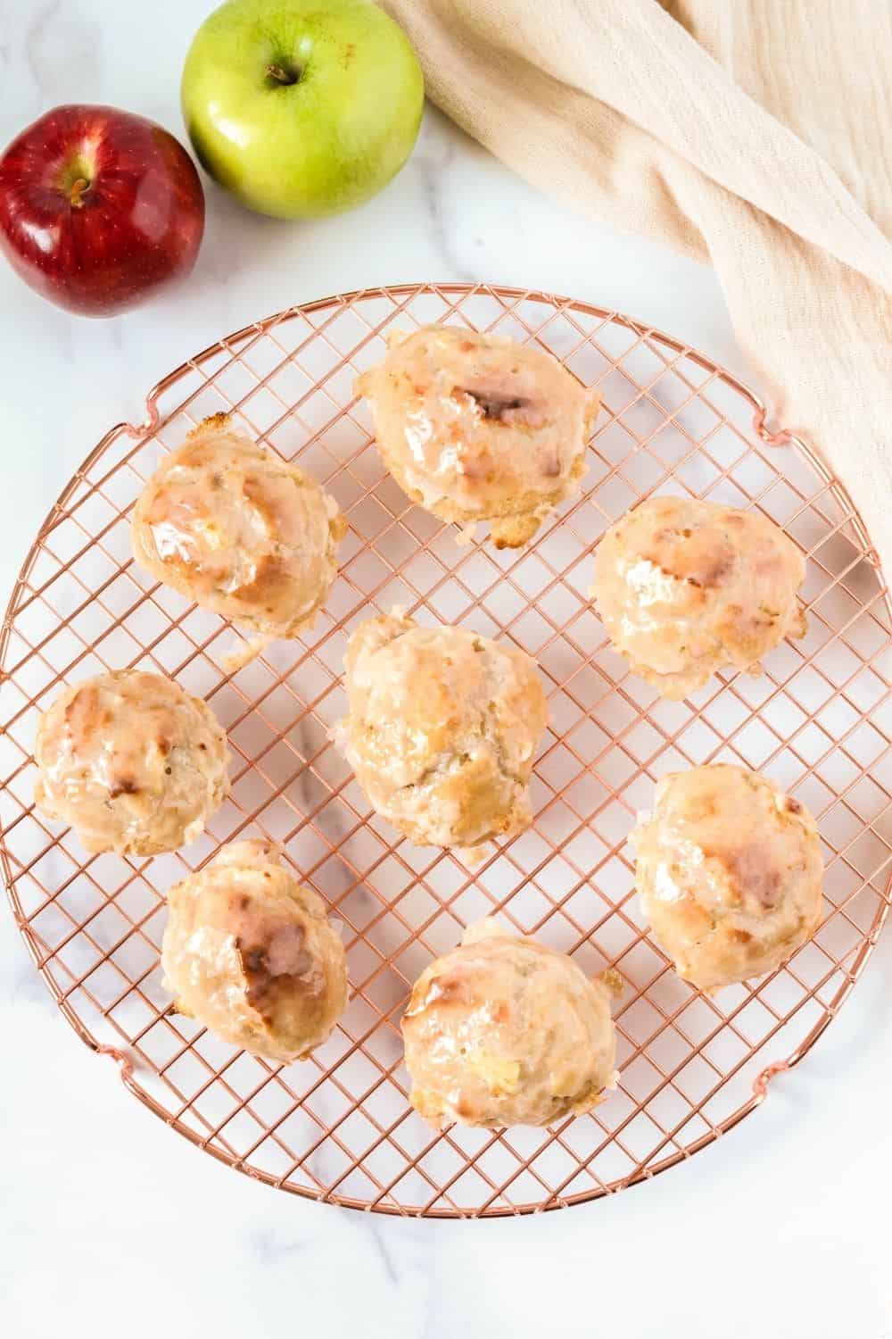 Overhead view of several baked apple fritters on a wire cooling rack. Two apples are next to the rack.