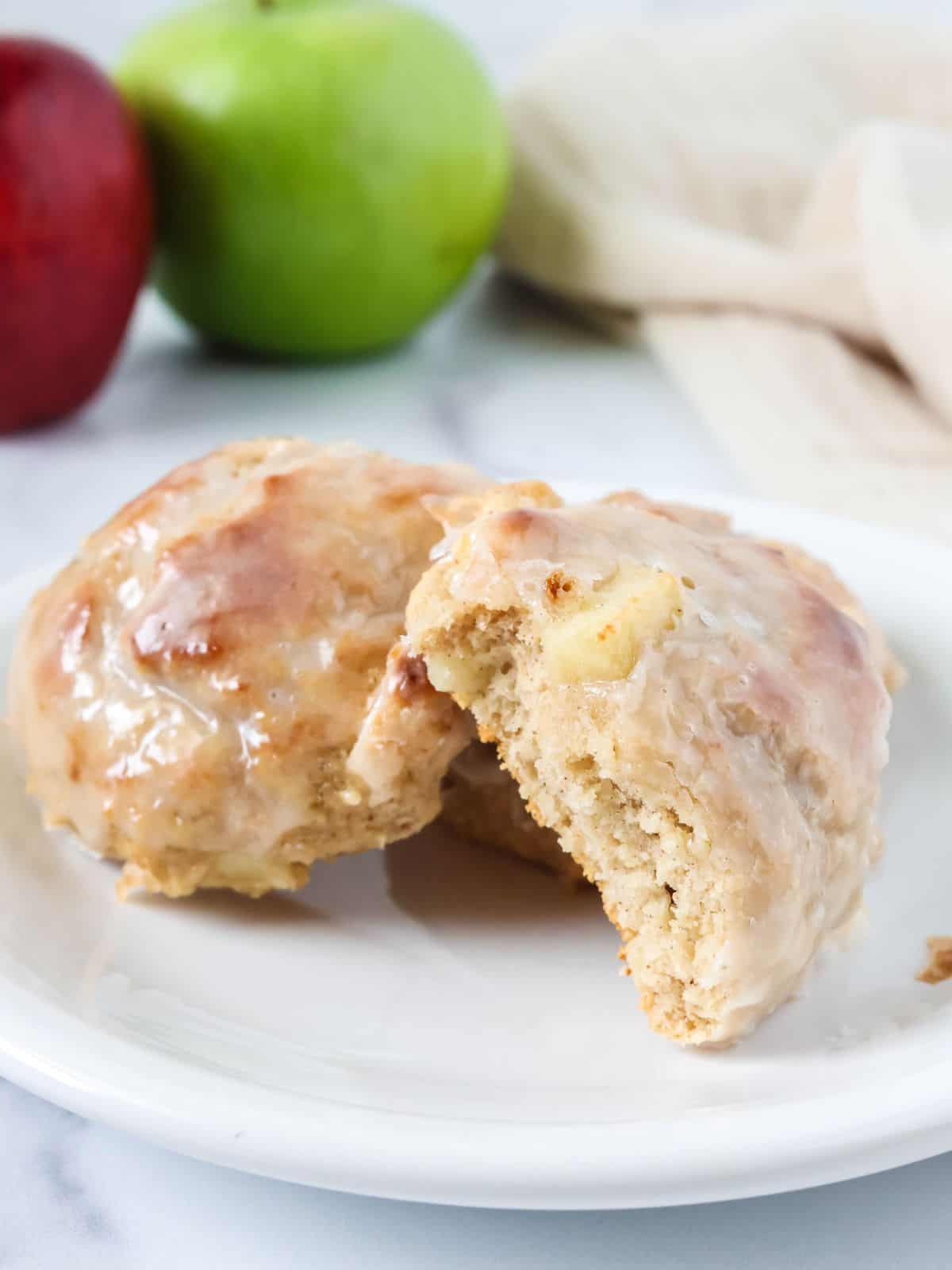 Baked apple fritters served on a white plate; one fritter has a bite taken out of it, showcasing the tender interior with chunks of apples.