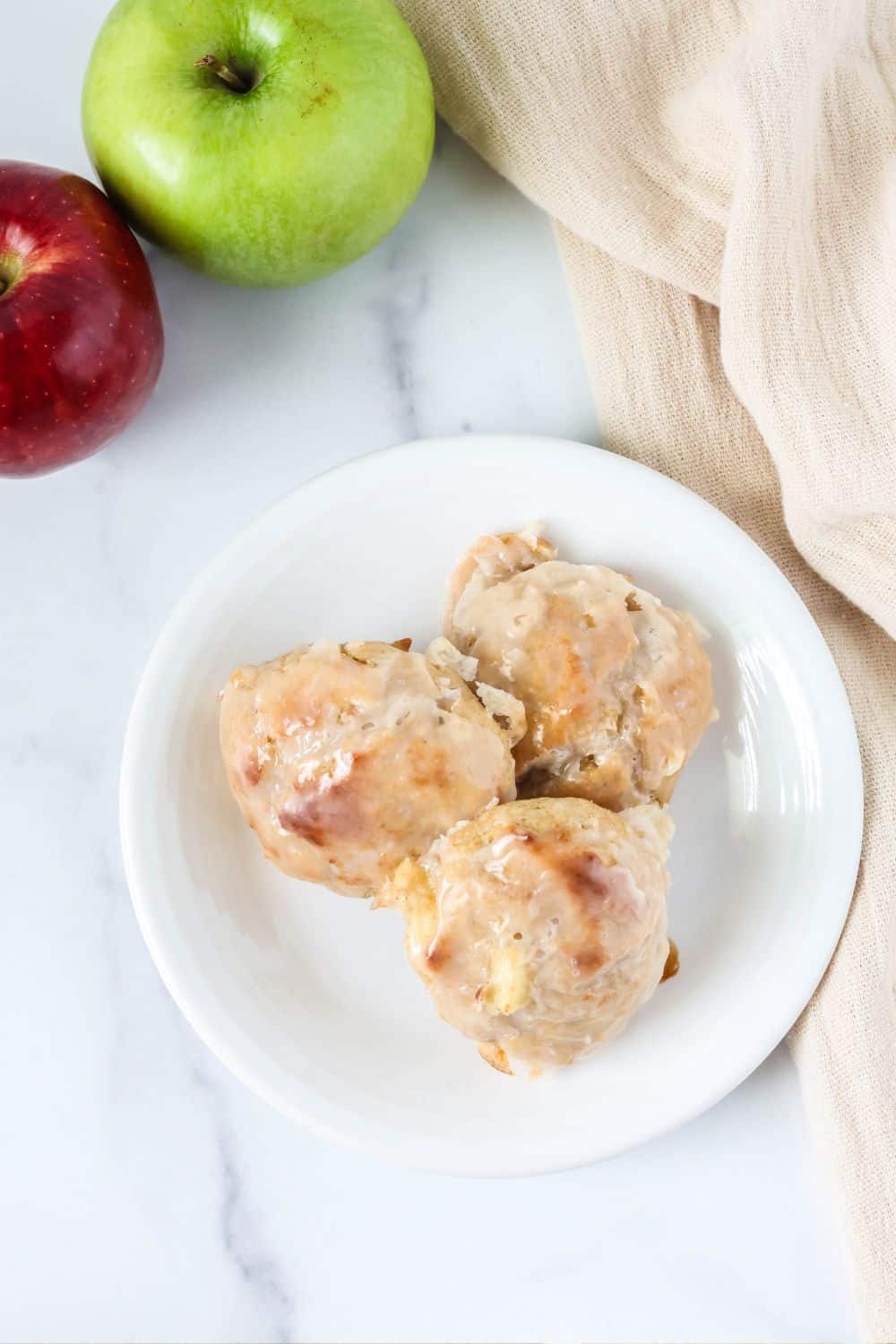 Overhead view of three baked apple fritters with glaze, served on a white plate. Two apples are in the background.