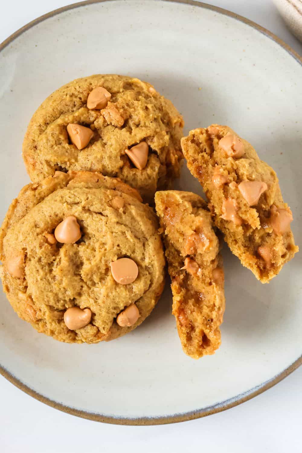 Overhead view of pumpkin butterscotch cookies on a plate; one cookie has been broken in half to show its tender interior and melty butterscotch chips.