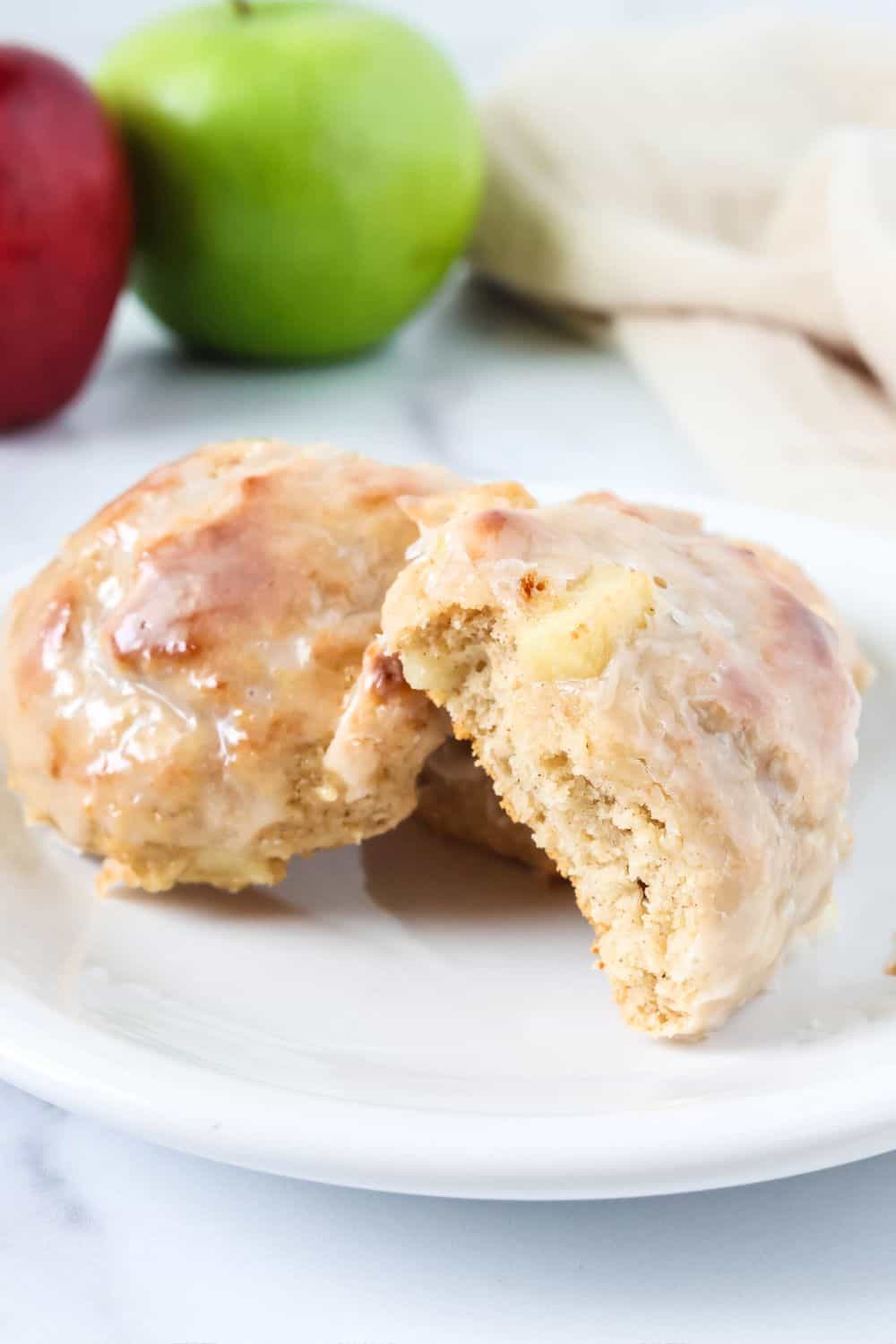 Fresh apple fritters, topped with glaze, on a white plate. One fritter has a bite taken out of it.