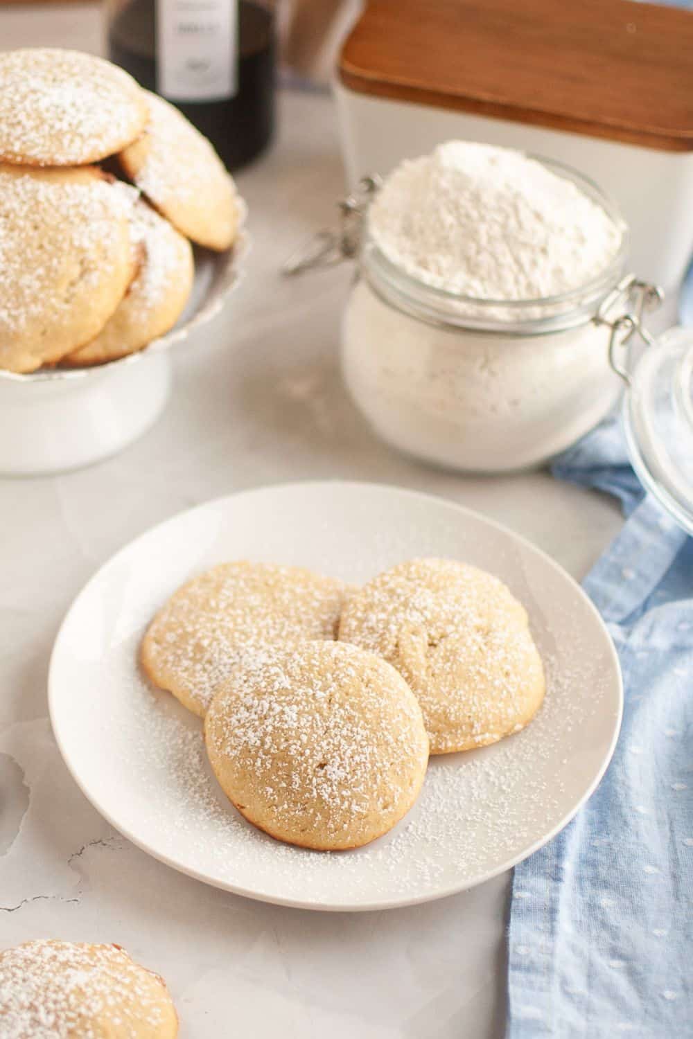 Three cream cheese cookies, dusted with powdered sugar, served on a white plate. More cookies and a canister of flour are in the background.