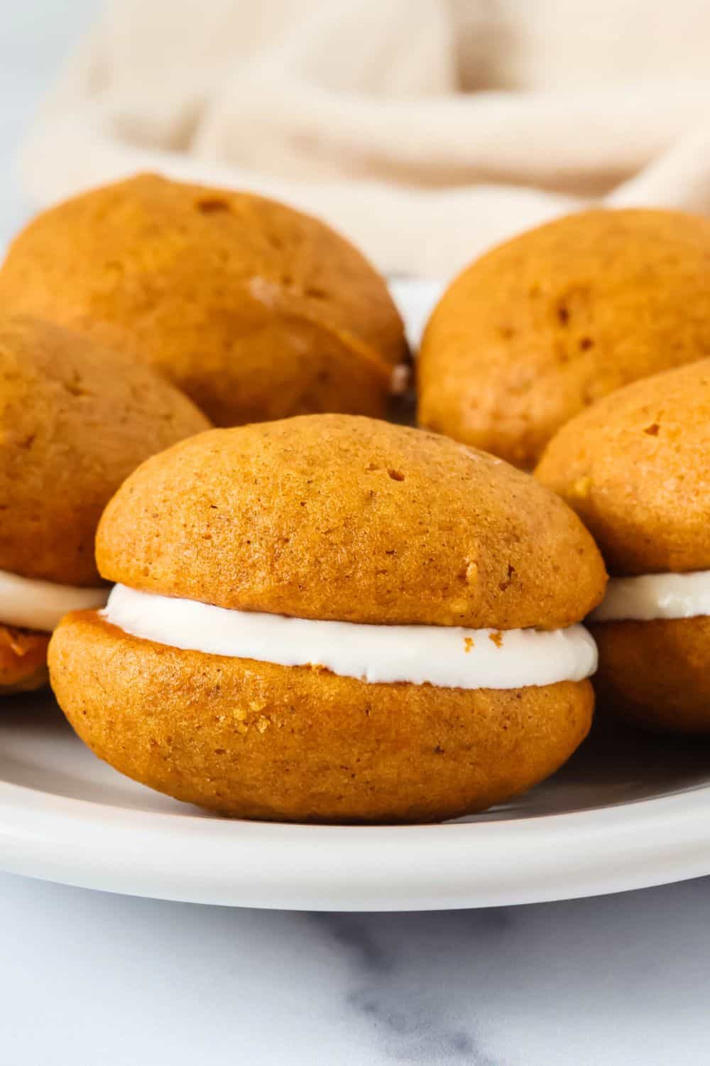 Close-up side view of fluffy pumpkin cream cheese cookie sandwiches on a white plate.