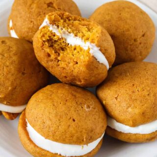 Pumpkin whoopie pies on a white plate. One cookie has a bite taken out of it to show the soft, cake-like texture of the cookies and the creamy marshmallow fluff filling.