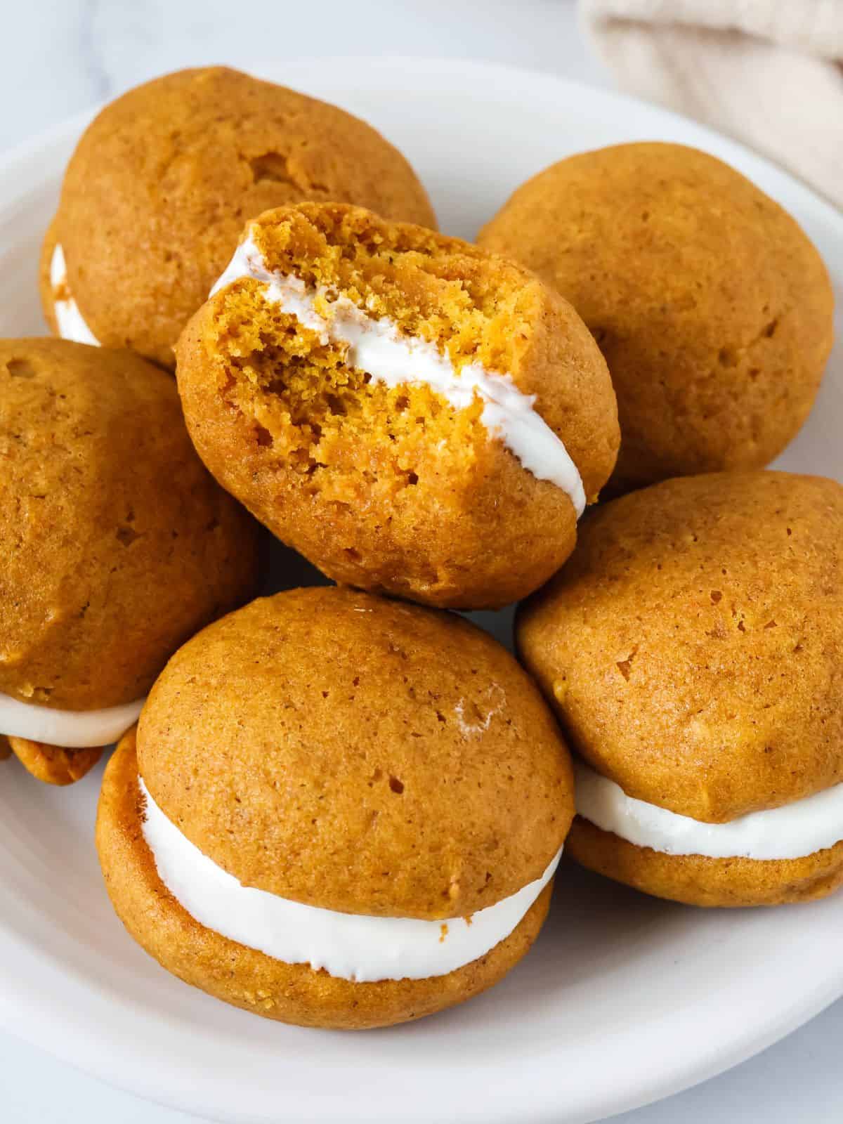 Pumpkin whoopie pies on a white plate. One cookie has a bite taken out of it to show the soft, cake-like texture of the cookies and the creamy marshmallow fluff filling.
