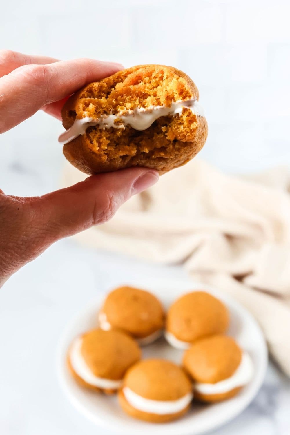 A hand holds a pumpkin whoopie pie with cream cheese marshmallow filling. A white plate of additional whoopie pies is in the background.