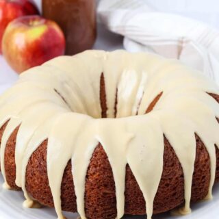 Apple butter bundt cake topped with brown sugar icing and served on a white platter. A jar of apple butter and two fresh apples are in the background.