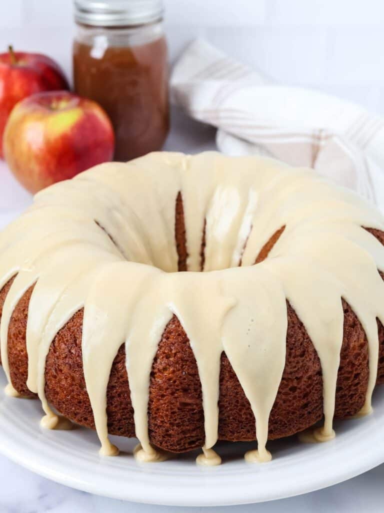 Apple butter bundt cake topped with brown sugar icing and served on a white platter. A jar of apple butter and two fresh apples are in the background.