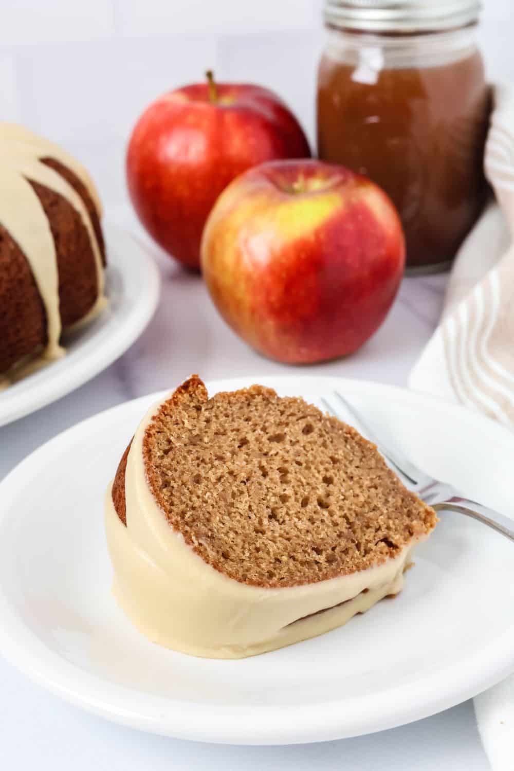 Close-up view of a slice of apple butter cake made from a cake mix, served on a white plate. In the background are a couple of fresh apples, a jar of apple butter, and the bundt cake.