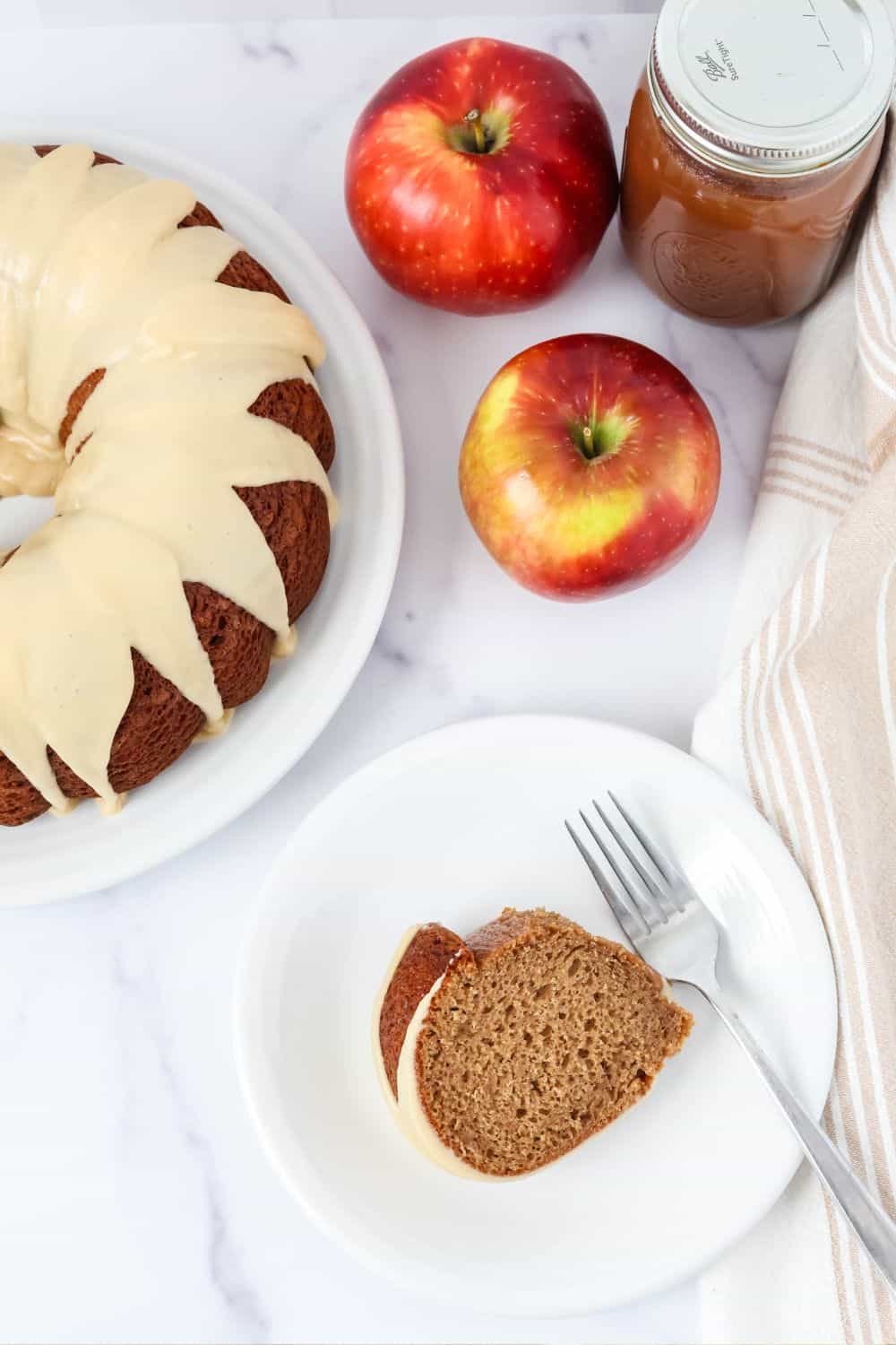 Overhead view of a slice of apple butter cake on a white dessert plate with a fork, a whole apple butter bundt cake topped with brown sugar icing on a white platter, two apples, and a jar of apple butter.