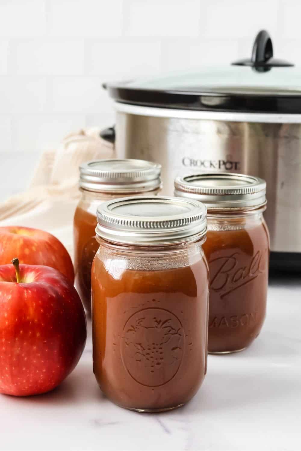 Three jars of homemade Amish-style apple butter in front of a Crock Pot slow cooker. Two apples are next to the jars.