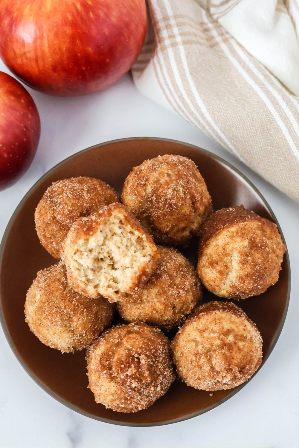 Overhead view of apple cider donut holes served on a brown plate, with fresh apples in the background. One of the donut holes has a bite taken out of it, showcasing the soft and tender interior.