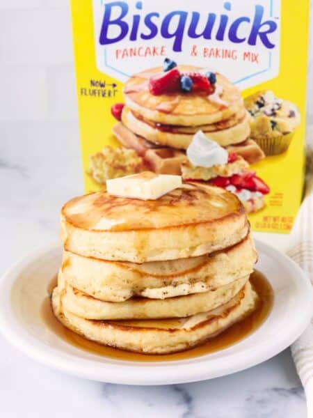 Stack of Bisquick pancakes on a white plate, topped with syrup and a pat of butter. A box of Bisquick pancake and baking mix is behind the plate of pancakes.