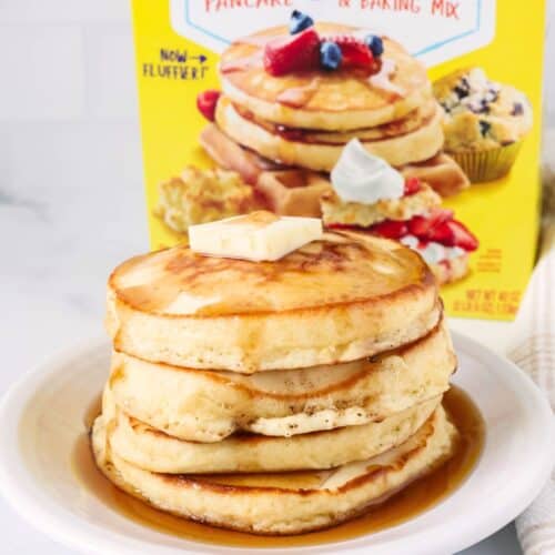 Stack of Bisquick pancakes on a white plate, topped with syrup and a pat of butter. A box of Bisquick pancake and baking mix is behind the plate of pancakes.