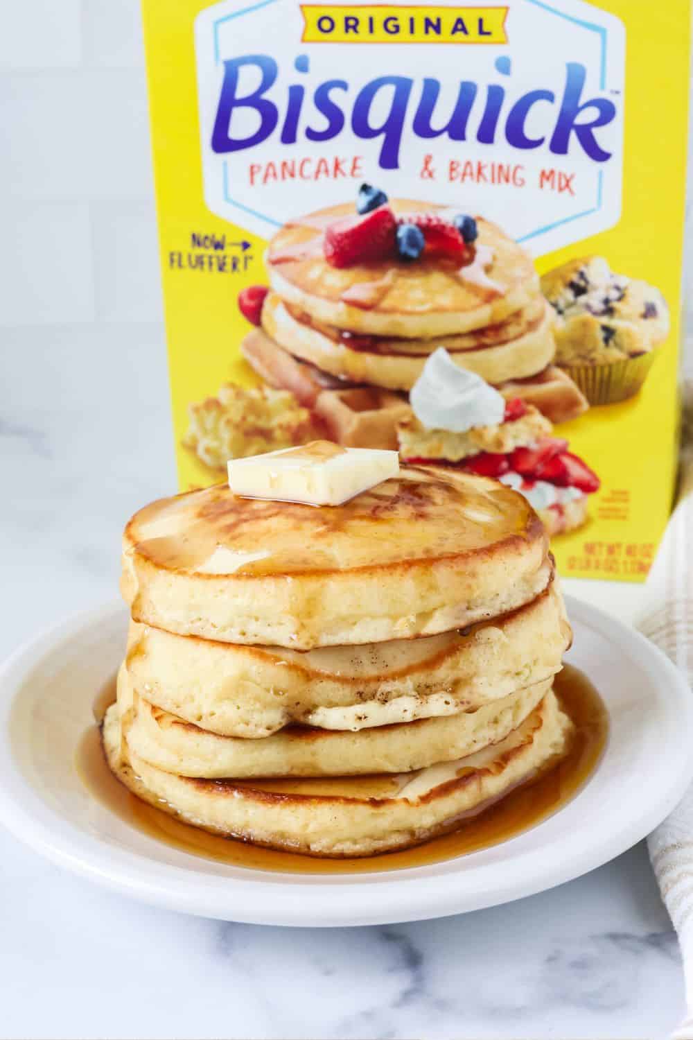 Stack of Bisquick pancakes on a white plate, topped with butter and syrup. A box of Bisquick pancake and baking mix is behind the plate.
