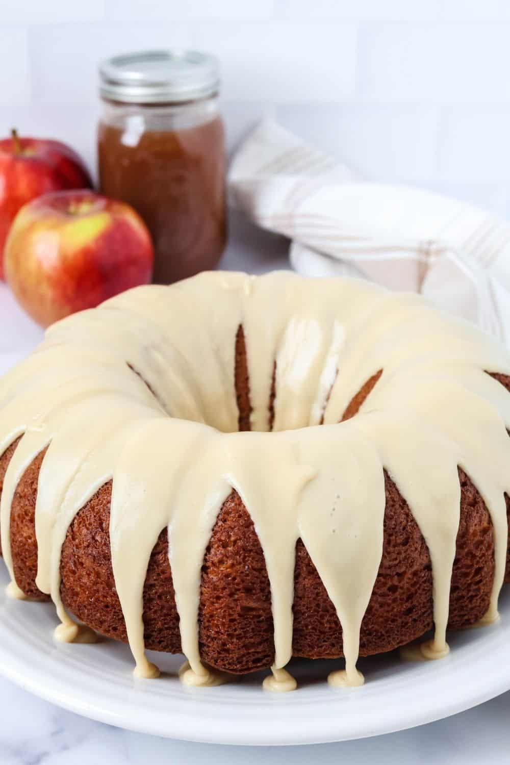 Apple butter bundt cake topped with brown sugar icing and served on a white plate. In the background are two apples and a jar of apple butter.