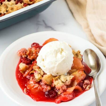 Cranberry apple crisp topped with a scoop of vanilla ice cream and served on a plate with a spoon. The baking dish of the fruit crisp is in the background.