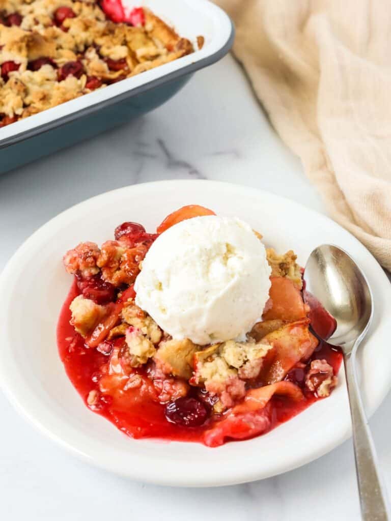 Cranberry apple crisp topped with a scoop of vanilla ice cream and served on a plate with a spoon. The baking dish of the fruit crisp is in the background.