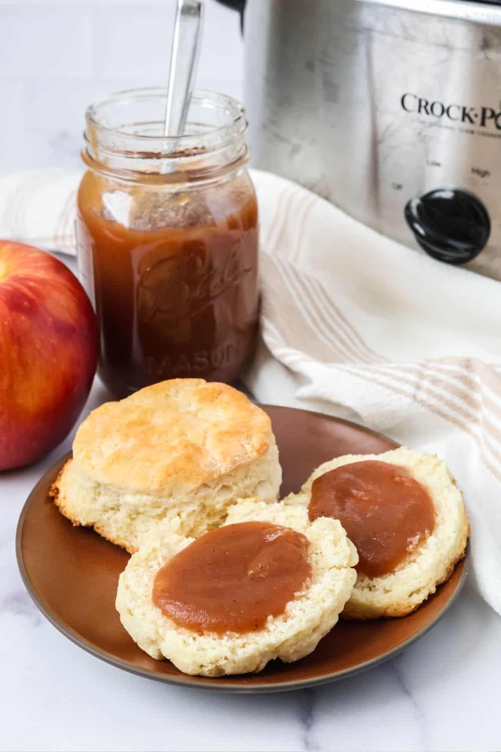 A brown plate of biscuits topped with apple butter. A jar of apple butter is in the background, alongside a Crock Pot slow cooker.