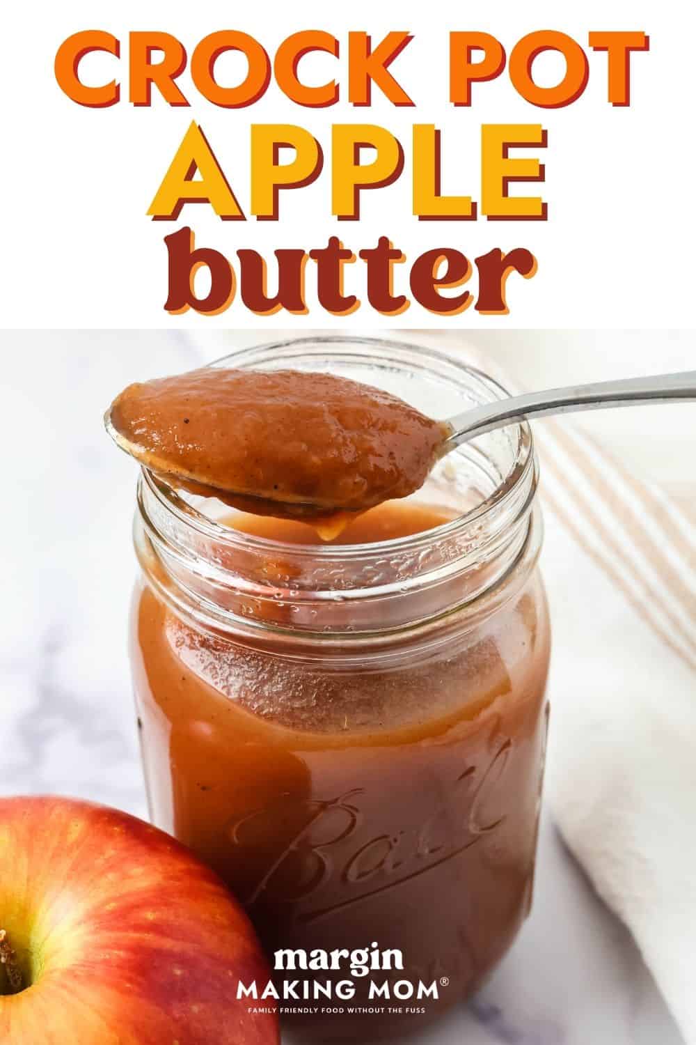 A spoonful of easy Crock Pot apple butter rests on the top of a jar of apple butter. A whole apple is next to the jar.