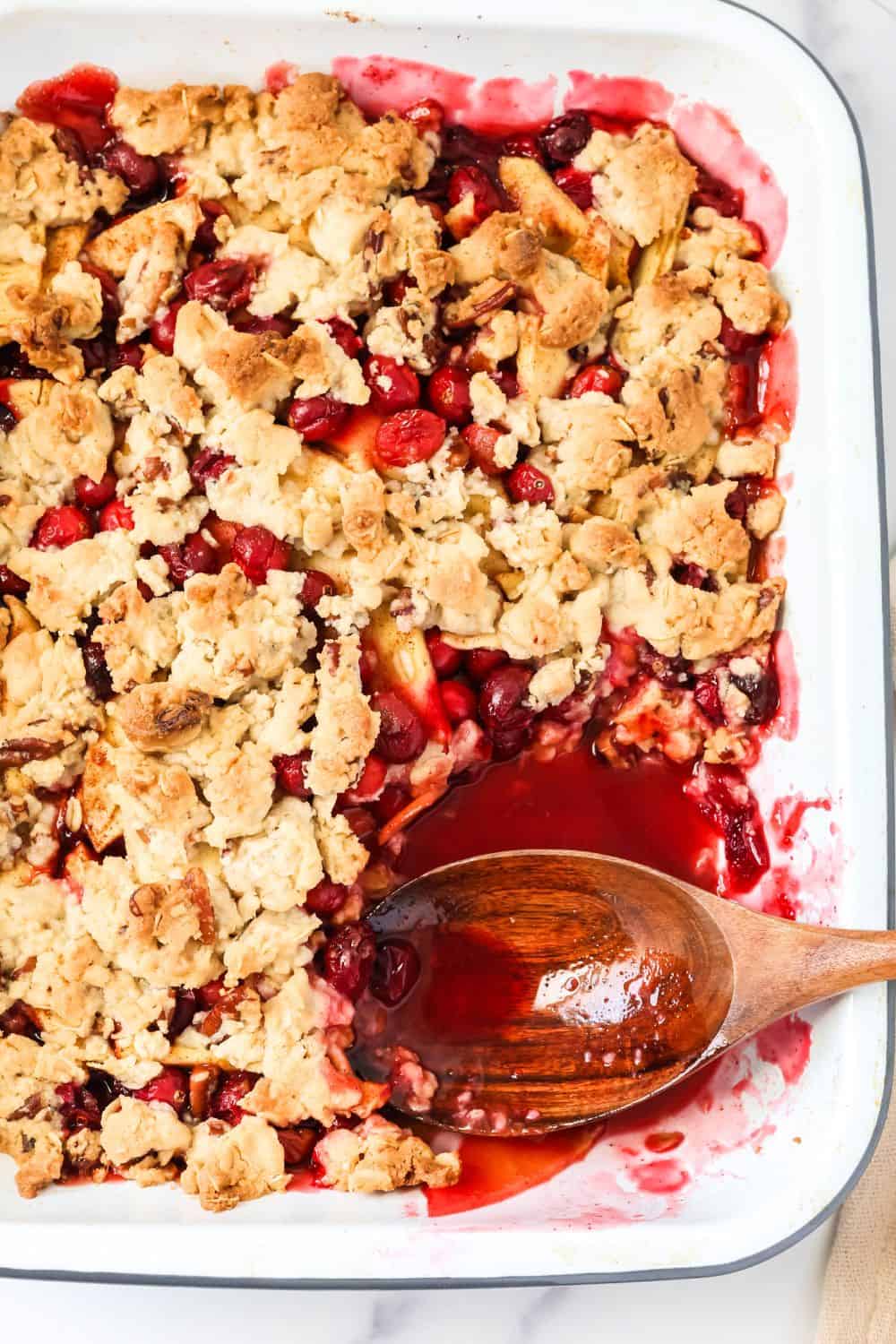 Baking dish of fall cranberry apple cobbler for Thanksgiving. A wooden serving spoon is in the dish, where a serving has been removed.