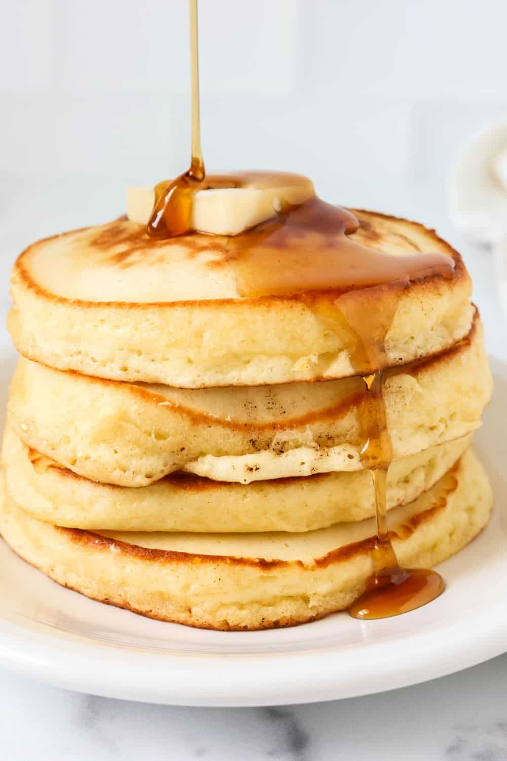 Syrup is being drizzled over a stack of fluffy Bisquick pancakes served on a white plate.