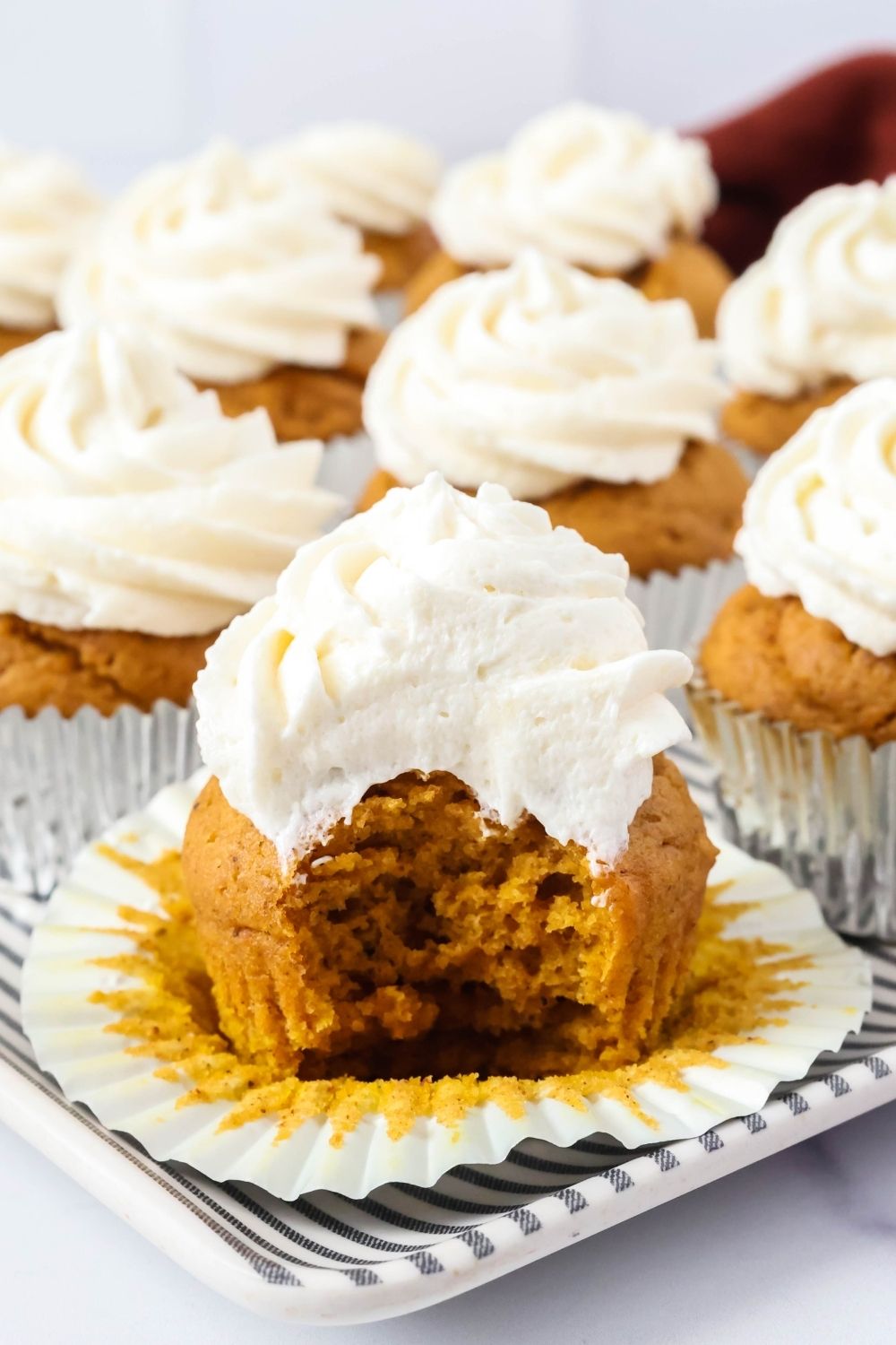 Several fluffy pumpkin cupcakes topped with ermine whipped cream frosting, served on a platter. The cupcake in the foreground has a bite taken out of it to show the moist texture inside.