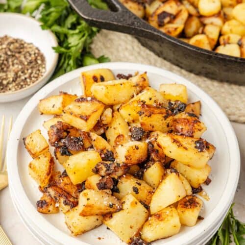 Fried potatoes and onions served on a white plate, with a cast iron skillet of more potatoes and onions in the background.