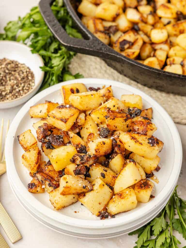 Fried potatoes and onions served on a white plate, with a cast iron skillet of more potatoes and onions in the background.