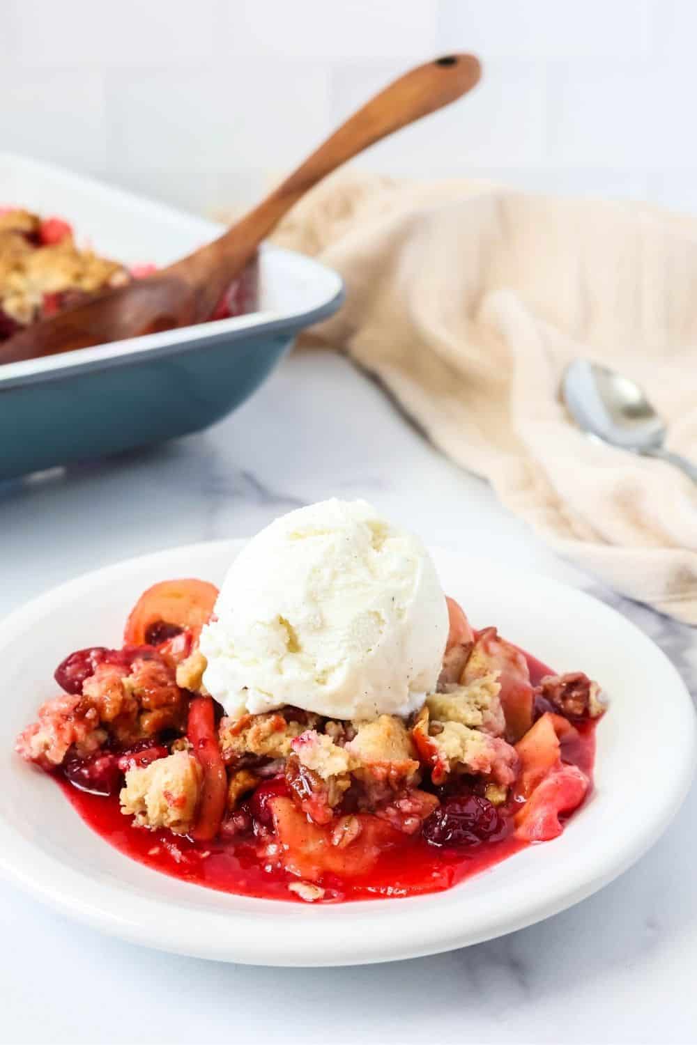 Homemade cranberry apple crisp on a white plate, topped with ice cream. The baking dish and serving spoon is in the background.