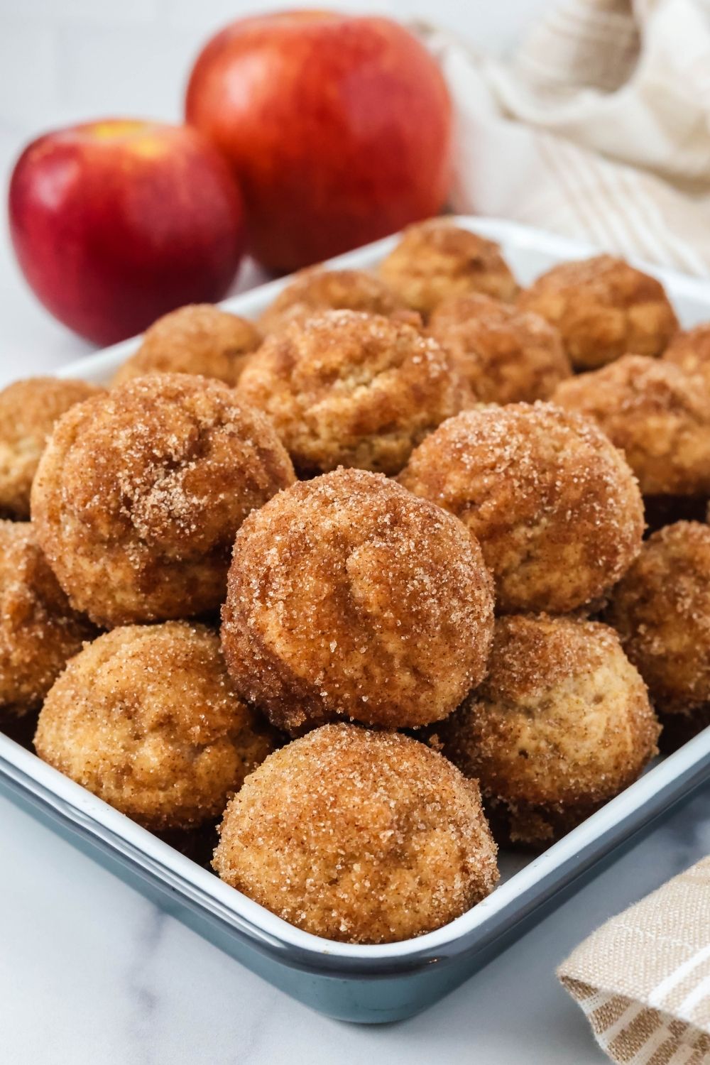 Several baked apple cider donut bites are served on a blue and white tray. Two apples are in the background.