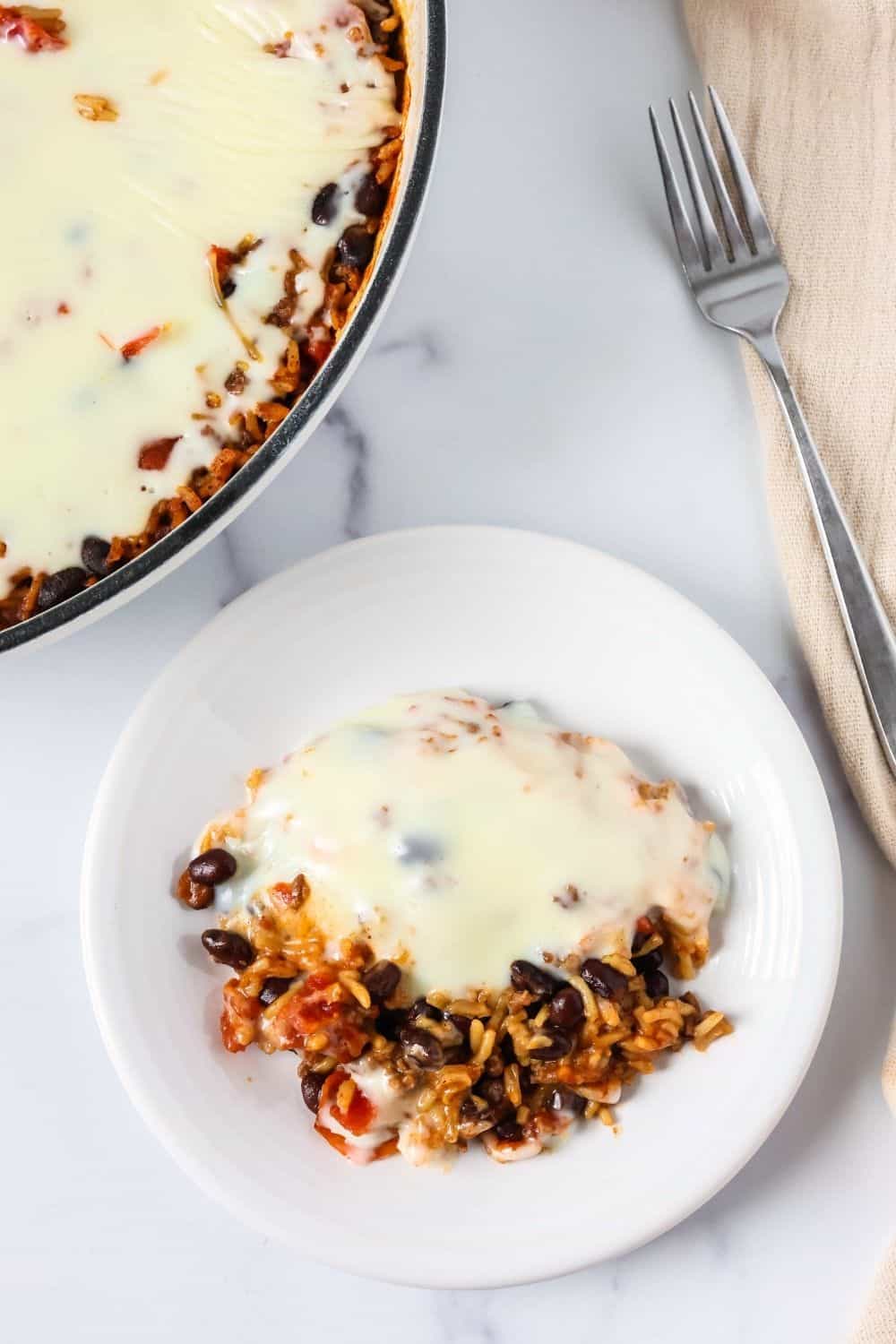 Overhead view of Knorr queso and rice casserole on a white plate, with the baking dish and a fork next to it.