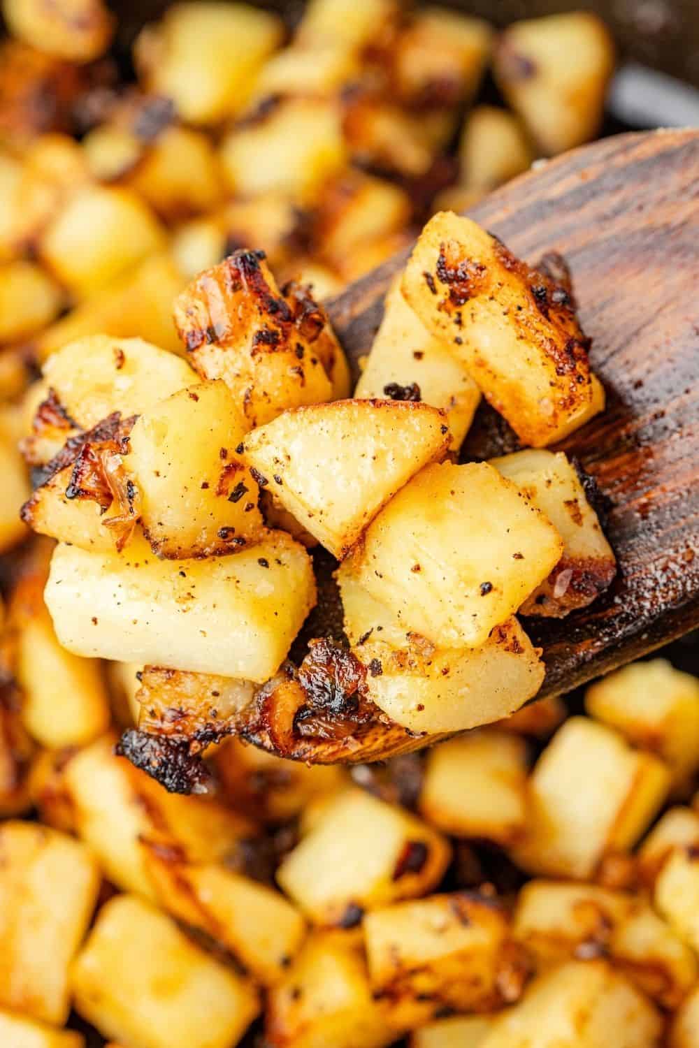 Close-up view of pan fried potatoes and onions, with a wooden spoon scooping out some of the golden-brown potatoes from the skillet.