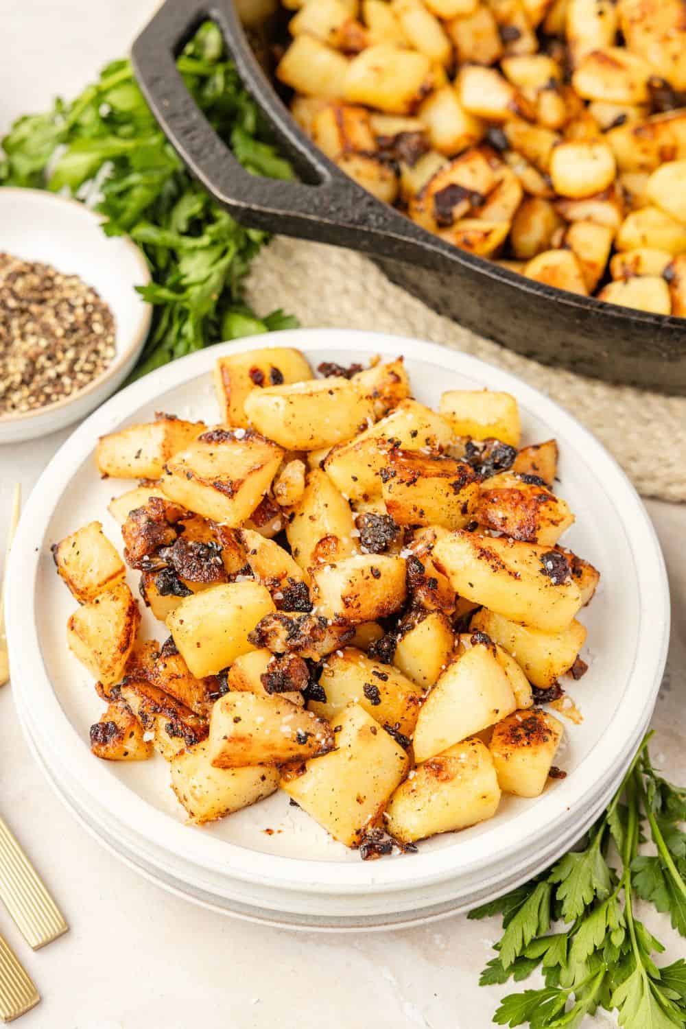 Potatoes and onions fried in bacon grease are served on a white plate. The cast iron skillet of remaining potatoes is in the background.