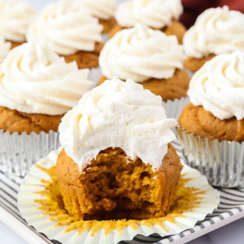 Several pumpkin cupcakes served on a platter; one cupcake has a bite taken out of it to show the texture of the cupcake and frosting.