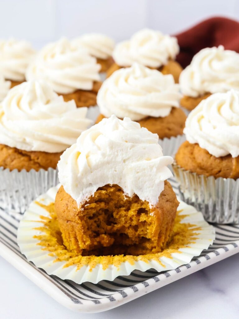 Several pumpkin cupcakes served on a platter; one cupcake has a bite taken out of it to show the texture of the cupcake and frosting.